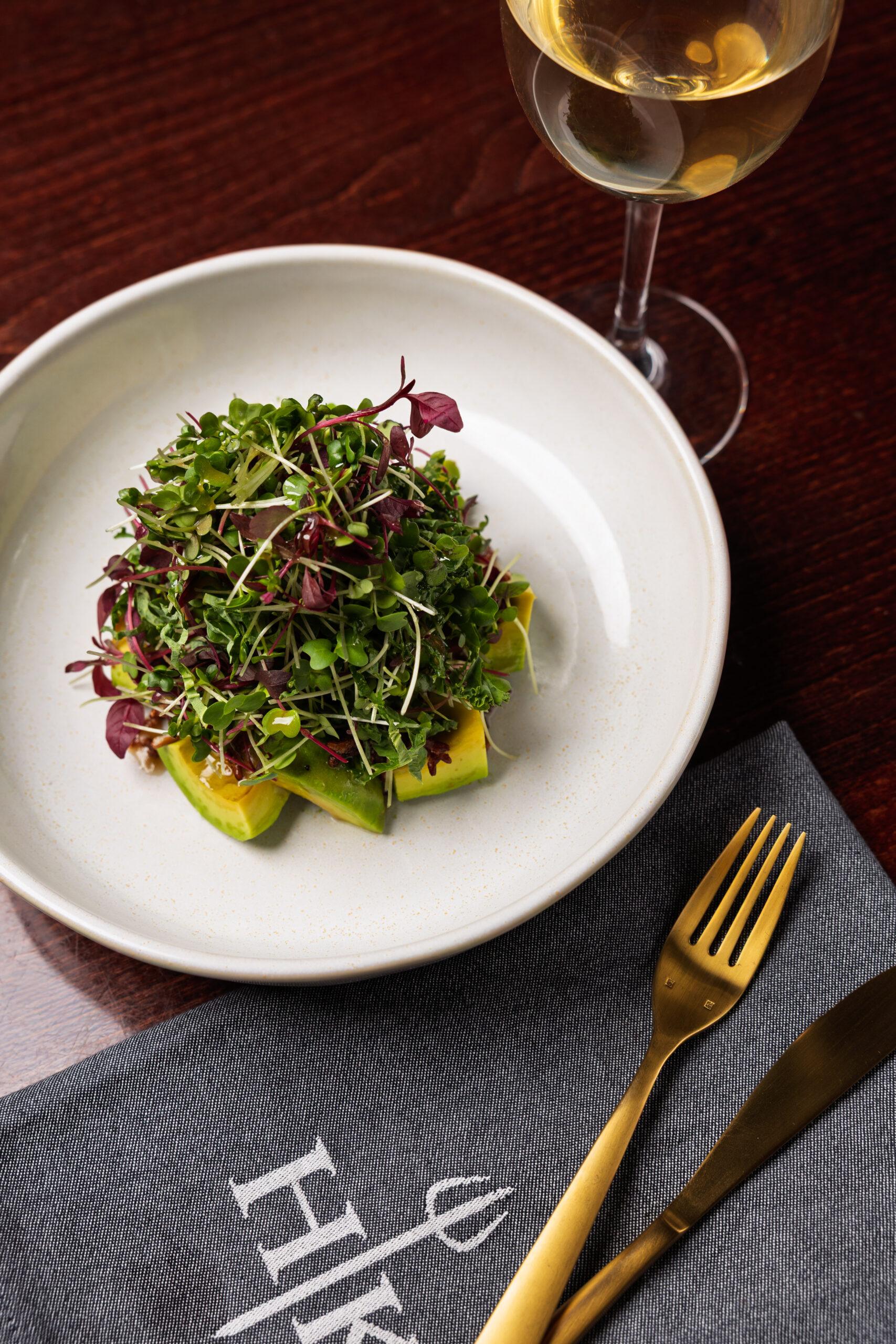 Avocado salad topped with mixed greens and herbs, served in a white bowl with a glass of white wine and gold cutlery on a restaurant table at Gordon Ramsay Hell’s Kitchen.