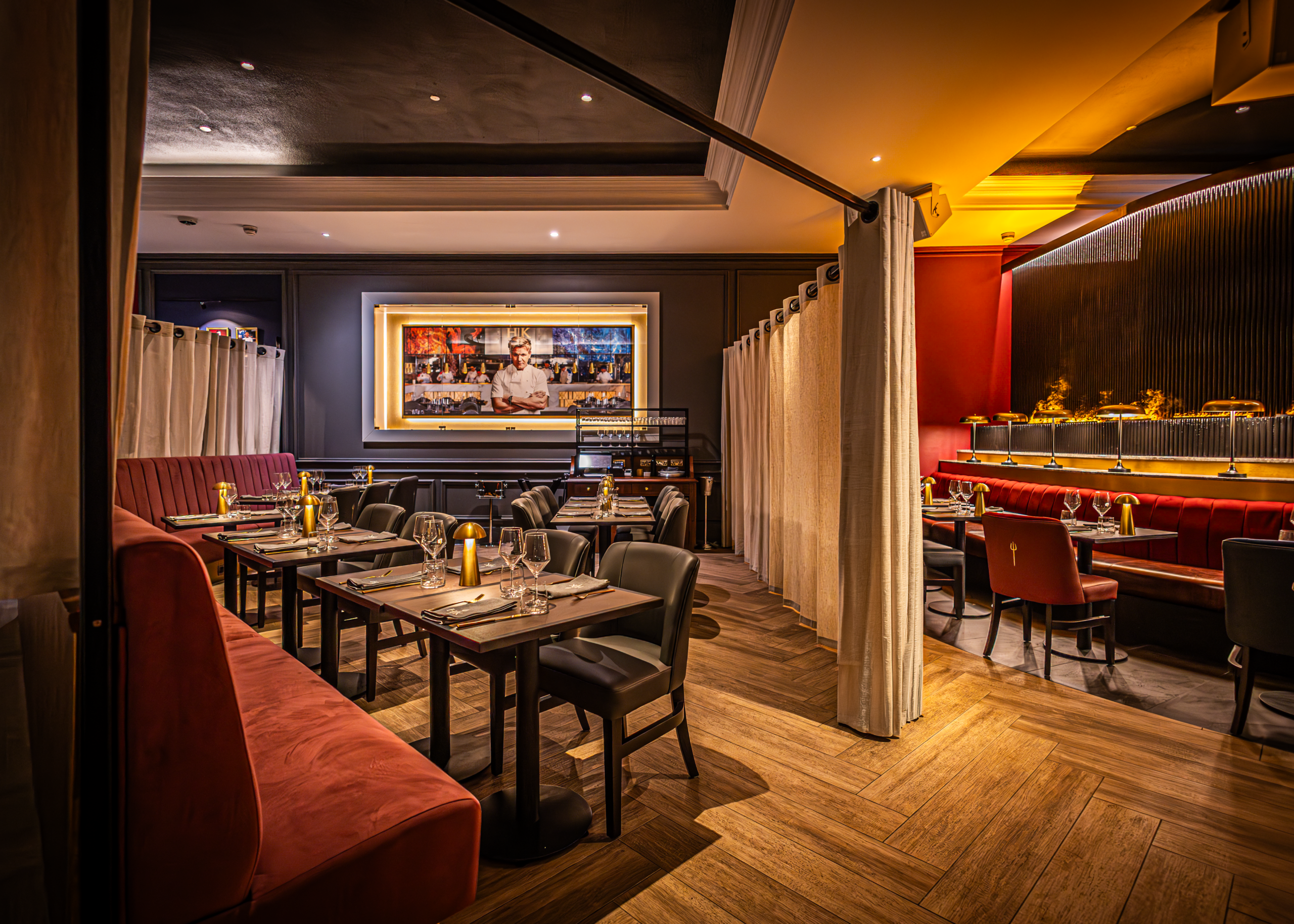 Interior of Hell's Kitchen London restaurant dining space with set tables, upholstered seating, warm ambient lighting, and curtain‑separated private dining area